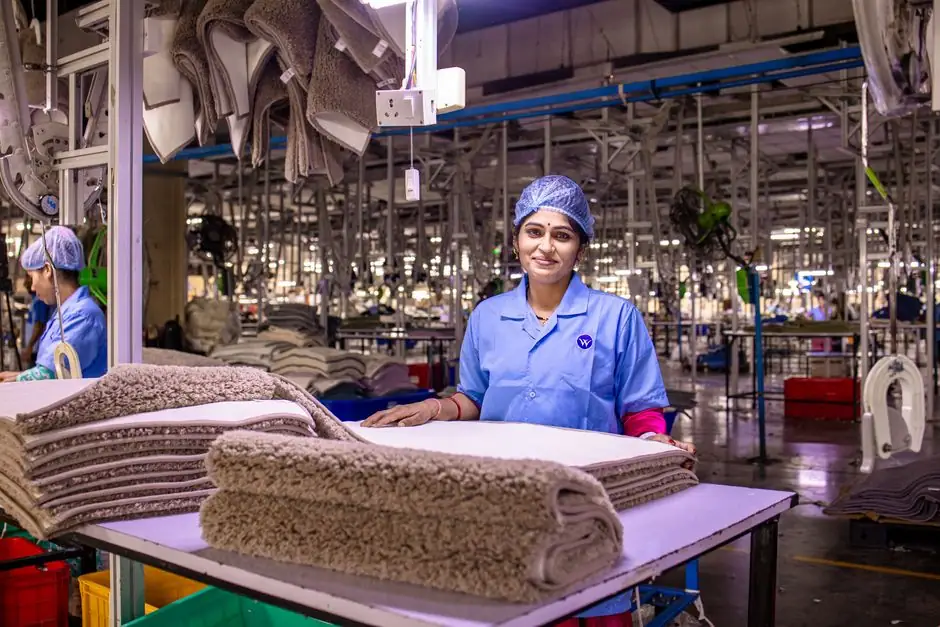 A female worker in a textile factory smiling, showcasing a stack of carpets.